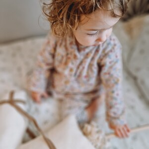Puede incluir: Una ni&ntilde;a peque&ntilde;a con el pelo rubio rizado se sienta en una cama blanca con almohadas blancas. La ni&ntilde;a lleva un mono de estampado floral rosa y blanco.