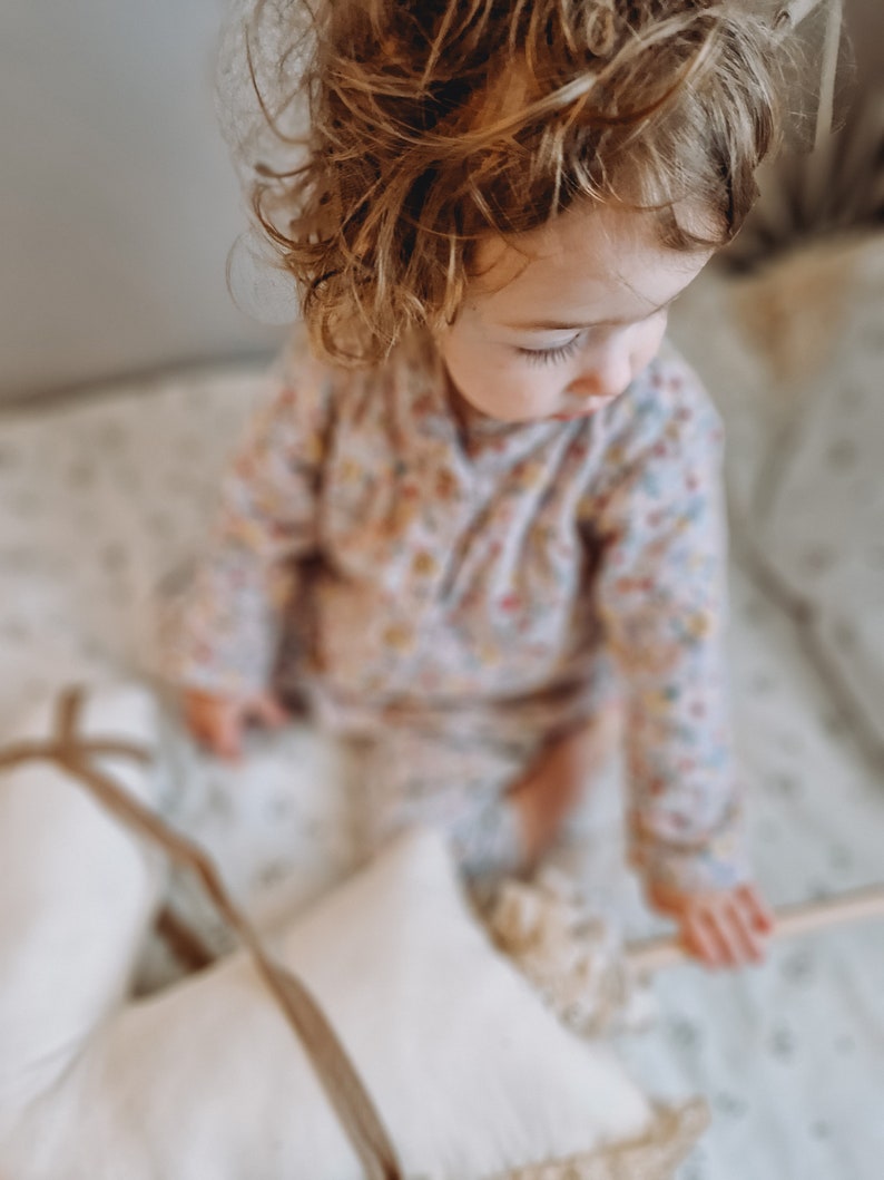 Puede incluir: Una ni&ntilde;a peque&ntilde;a con el pelo rubio rizado se sienta en una cama blanca con almohadas blancas. La ni&ntilde;a lleva un mono de estampado floral rosa y blanco.