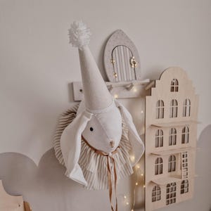 Könnte beinhalten: Ein weißer Stoffhasenkopf mit einem weißen und braunen gestreiften Rüschenkragen und einer weißen Partymütze mit einem weißen Bommel oben. Der Hasenkopf ist an einer Wand mit einem Holzhaus und einer kleinen Holztür dahinter befestigt.
