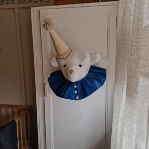 Può includere: Una testa di orso di peluche bianco con un colletto a quadri blu e bianco e un cappello da festa dorato e bianco. La testa di orso è montata su una porta bianca.
