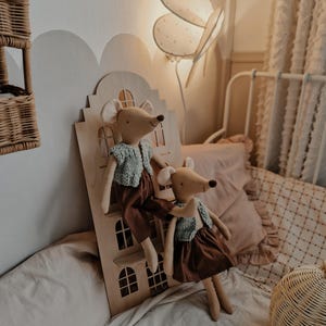 May include: Two brown plush mice with knitted green sweaters and brown skirts are standing on a wooden dollhouse. The dollhouse has a white butterfly lamp on the wall behind it.