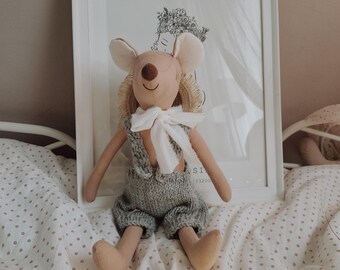 May include: A handmade plush mouse doll with a tan body, large ears, and gray overalls. The doll has a white bow tie and is sitting on a bed with a white framed picture in the background. The picture has a black and white illustration.