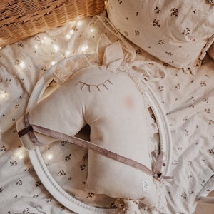 Puede incluir: Una cabeza de caballo de peluche blanca con una cinta rosa y un borde de encaje blanco. La cabeza de caballo est&aacute; sobre un fondo blanco con un patr&oacute;n floral.