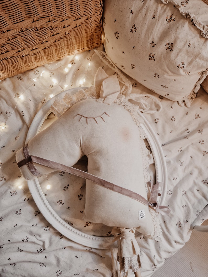Puede incluir: Una cabeza de caballo de peluche blanca con una cinta rosa y un borde de encaje blanco. La cabeza de caballo est&aacute; sobre un fondo blanco con un patr&oacute;n floral.
