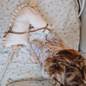 Puede incluir: Una ni&ntilde;a peque&ntilde;a est&aacute; sentada en una cama con una s&aacute;bana blanca con estampado floral, sosteniendo un caballo de balanc&iacute;n de peluche blanco con riendas marrones. La ni&ntilde;a lleva una camiseta de pijama con estampado floral y tiene el pelo largo y rizado marr&oacute;n.