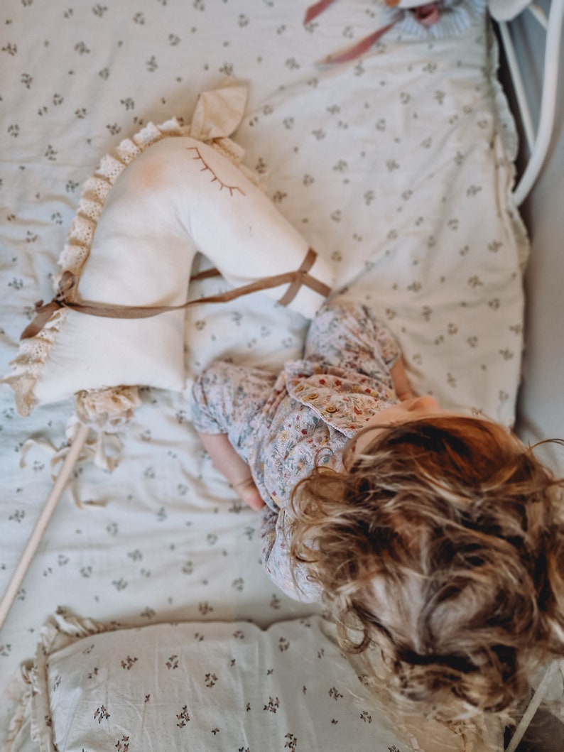 Puede incluir: Una ni&ntilde;a peque&ntilde;a est&aacute; sentada en una cama con una s&aacute;bana blanca con estampado floral, sosteniendo un caballo de balanc&iacute;n de peluche blanco con riendas marrones. La ni&ntilde;a lleva una camiseta de pijama con estampado floral y tiene el pelo largo y rizado marr&oacute;n.