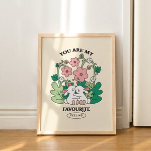 May include: A framed print with a light beige background featuring two white rabbits hugging in a field of green leaves and pink flowers. The text "You are my favourite feeling" is printed below the rabbits.