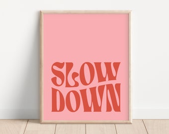 May include: A pink framed print with the text "Slow Down" in red lettering. The text is in a retro font style.