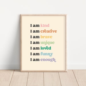 Op de afbeelding: Een ingelijst print met een lichtgele achtergrond en zwarte tekst die "Ik ben aardig", "Ik ben creatief", "Ik ben dapper", "Ik ben uniek", "Ik ben geliefd", "Ik ben grappig" en "Ik ben genoeg" luidt. De woorden zijn in verschillende kleuren, waaronder roze, oranje, geel, groen, blauw en paars.