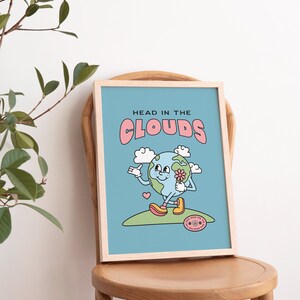 May include: A light blue framed print with the text "HEAD IN THE CLOUDS" in pink letters. The print features a cartoon illustration of the Earth with a smiling face, holding a flower and walking on a green hill. The Earth is wearing yellow shoes and has a pink heart above it. The print also includes a small circular sticker with the text "GOOD VIBES FOR THE VIBES YOU GIVE" in pink letters.
