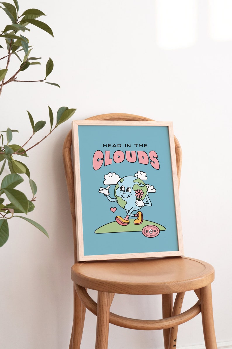 May include: A light blue framed print with the text "HEAD IN THE CLOUDS" in pink letters. The print features a cartoon illustration of the Earth with a smiling face, holding a flower and walking on a green hill. The Earth is wearing yellow shoes and has a pink heart above it. The print also includes a small circular sticker with the text "GOOD VIBES FOR THE VIBES YOU GIVE" in pink letters.