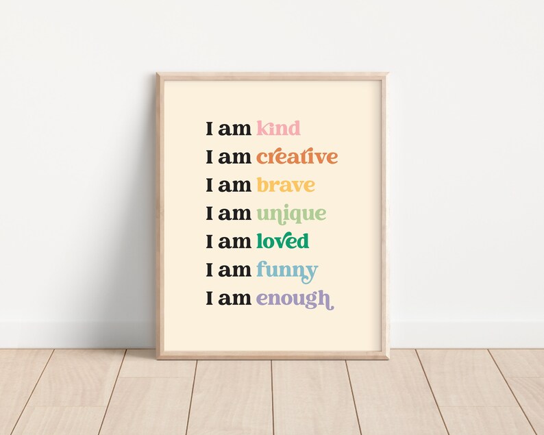 Op de afbeelding: Een ingelijst print met een lichtgele achtergrond en zwarte tekst die "Ik ben aardig", "Ik ben creatief", "Ik ben dapper", "Ik ben uniek", "Ik ben geliefd", "Ik ben grappig" en "Ik ben genoeg" luidt. De woorden zijn in verschillende kleuren, waaronder roze, oranje, geel, groen, blauw en paars.