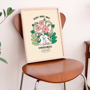 May include: A framed print with a light yellow background featuring two white bunnies hugging in a field of pink and white flowers. The text "You are my favourite feeling" is printed in black below the bunnies.
