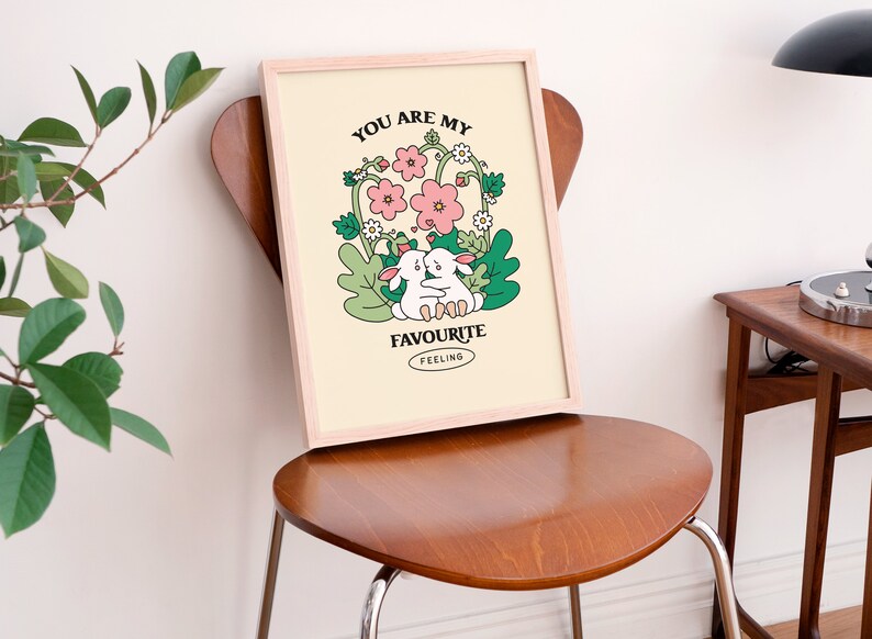 May include: A framed print with a light yellow background featuring two white bunnies hugging in a field of pink and white flowers. The text "You are my favourite feeling" is printed in black below the bunnies.