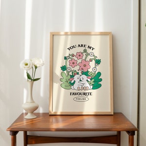 May include: A framed print with a light beige background featuring two white rabbits hugging in a field of green leaves and pink flowers. The text "You are my favourite feeling" is printed below the rabbits.