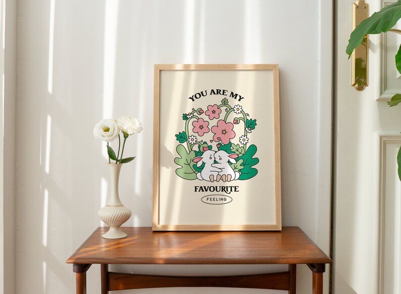 May include: A framed print with a light beige background featuring two white rabbits hugging in a field of green leaves and pink flowers. The text "You are my favourite feeling" is printed below the rabbits.