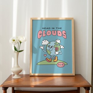 May include: A framed print with a blue background and a cartoon illustration of a smiling earth holding a flower. The text "HEAD IN THE CLOUDS" is in pink letters above the earth.