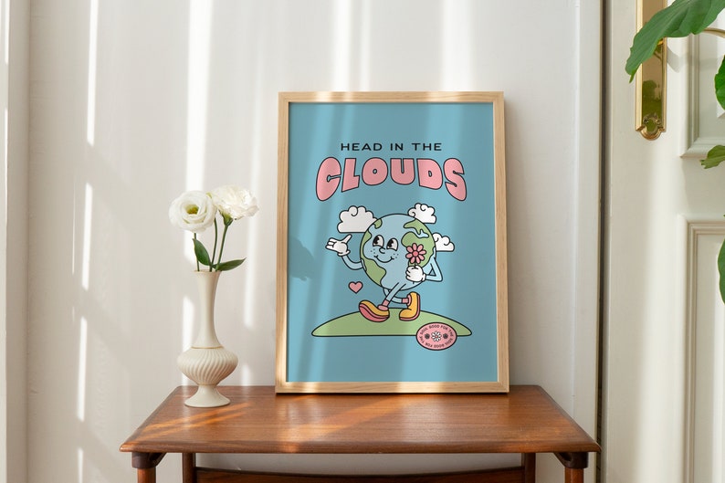 May include: A framed print with a blue background and a cartoon illustration of a smiling earth holding a flower. The text "HEAD IN THE CLOUDS" is in pink letters above the earth.