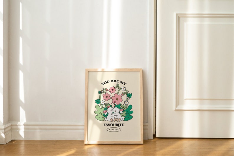 May include: A framed print with a light beige background featuring two white rabbits hugging in a field of green leaves and pink flowers. The text "You are my favourite feeling" is printed below the rabbits.