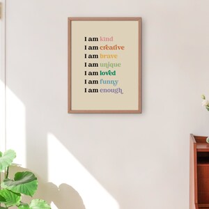 Op de afbeelding: Een ingelijst print met een beige achtergrond en zwarte tekst die "Ik ben aardig", "Ik ben creatief", "Ik ben dapper", "Ik ben uniek", "Ik ben geliefd", "Ik ben grappig" en "Ik ben genoeg" luidt. De tekst is in verschillende kleuren, waaronder roze, oranje, geel, groen, blauw en paars.