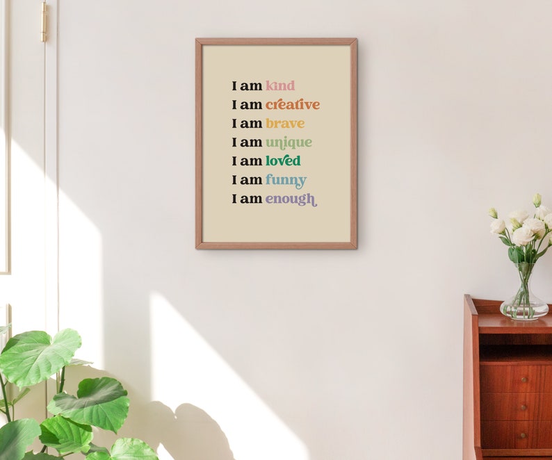 Op de afbeelding: Een ingelijst print met een beige achtergrond en zwarte tekst die "Ik ben aardig", "Ik ben creatief", "Ik ben dapper", "Ik ben uniek", "Ik ben geliefd", "Ik ben grappig" en "Ik ben genoeg" luidt. De tekst is in verschillende kleuren, waaronder roze, oranje, geel, groen, blauw en paars.