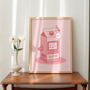 May include: A framed print with a pink background featuring a cartoon illustration of a pink carton of "Self Love Juice" with a red heart and the text "Fresh!" in a white circle. The carton has a barcode and three red hearts on the front.