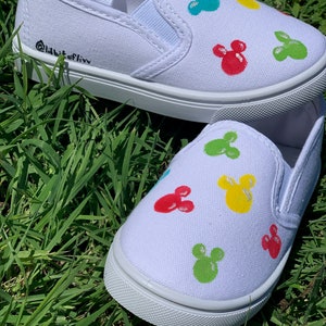 Może przedstawiać: Białe pł&oacute;cienne buty typu slip-on z kolorowym wzorem Myszki Miki. Buty mają czarną etykietę tekstową z napisem "Addicted to Liv".