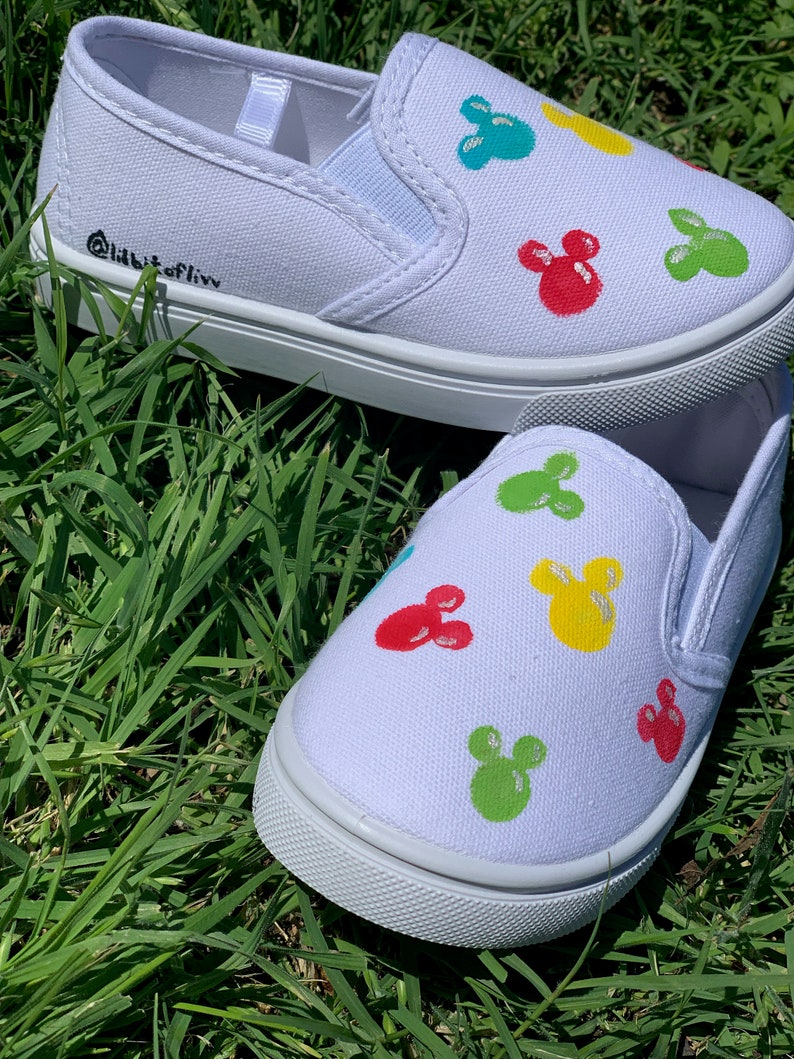 Może przedstawiać: Białe pł&oacute;cienne buty typu slip-on z kolorowym wzorem Myszki Miki. Buty mają czarną etykietę tekstową z napisem "Addicted to Liv".