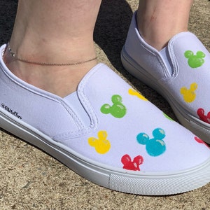 Może przedstawiać: Białe pł&oacute;cienne buty typu slip-on z kolorowym wzorem Myszki Miki. Buty mają białą gumową podeszwę i są ozdobione czerwonymi, ż&oacute;łtymi, zielonymi i niebieskimi głowami Myszki Miki.