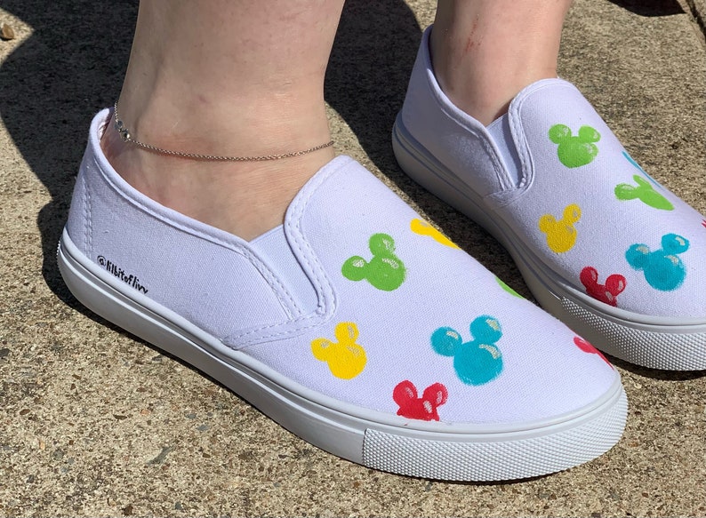 Może przedstawiać: Białe pł&oacute;cienne buty typu slip-on z kolorowym wzorem Myszki Miki. Buty mają białą gumową podeszwę i są ozdobione czerwonymi, ż&oacute;łtymi, zielonymi i niebieskimi głowami Myszki Miki.