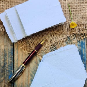 May include: A stack of handmade, light blue paper with deckled edges, alongside a matching envelope set. A burgundy and gold pen rests nearby on a rustic wooden surface, with a burlap cloth and yellow flowers in the background.