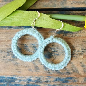 May include: A pair of light blue, crocheted hoop earrings with silver-colored hooks. The earrings are round and fluffy, resting on a wooden surface with a green leaf and a yellow tulip.