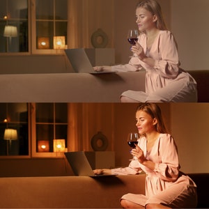 May include: A woman sits on a couch in a dimly lit room, holding a glass of red wine and using a laptop. She is wearing a light pink dress.