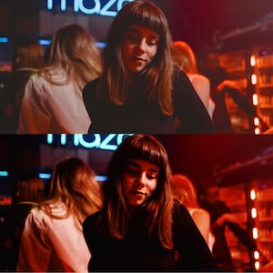 May include: Two women with long dark hair are standing in a dimly lit bar. The woman in the foreground is wearing a black turtleneck sweater and the woman in the background is wearing a white shirt. The bar is decorated with neon lights and there are other people in the background.