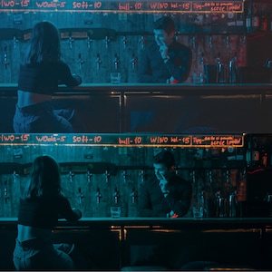 May include: Two people sitting at a bar counter. The woman is wearing a black crop top and blue jeans. The man is wearing a black shirt and blue jeans. The bar counter has a sign with the words "T-10 Plwo-15 Woola-5 soft-10 bull-10 WINO kel-15 Tipy-Steelled" written on it.