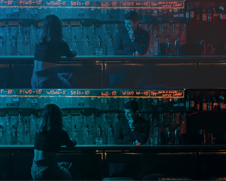 May include: Two people sitting at a bar counter. The woman is wearing a black crop top and blue jeans. The man is wearing a black shirt and blue jeans. The bar counter has a sign with the words "T-10 Plwo-15 Woola-5 soft-10 bull-10 WINO kel-15 Tipy-Steelled" written on it.