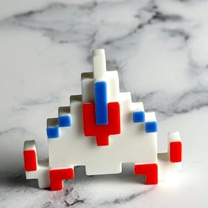May include: A white, pixelated spaceship pin with red and blue details. The pin is a retro design, reminiscent of classic arcade games. The spaceship has a triangular shape with a red centre and blue details.