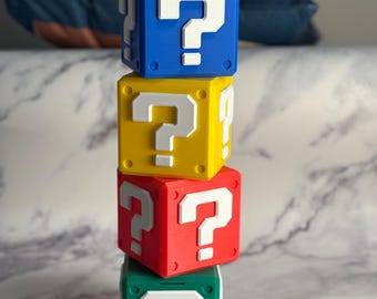 May include: Four stacked, colorful question mark blocks. The top block is blue, the second is yellow, the third is red, and the bottom block is green. Each block has a white question mark on the front.
