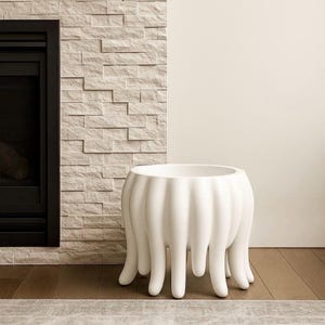 May include: A white, modern planter with a unique design. The planter has a rounded body with vertical ridges and elongated, tentacle-like legs. It is placed on a wooden floor, with a textured stone wall and fireplace in the background.