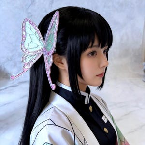 May include: A large butterfly hair clip in shades of pink, green, and white is attached to black hair. The clip is worn on the side of the head. The person is wearing a black and white outfit with gold buttons.