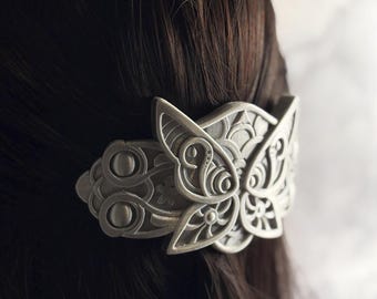 May include: A silver-colored hair clip with an ornate butterfly design. The clip is made of metal and features detailed floral and geometric patterns. The butterfly wings are open, and the clip is attached to dark brown hair.