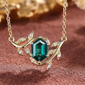 Puede incluir: Un collar dorado con una piedra preciosa central de color verde oscuro, talla esmeralda. El colgante está enmarcado por delicados diseños en forma de hojas adornados con pequeños acentos brillantes. La fina cadena complementa la elegancia del colgante.