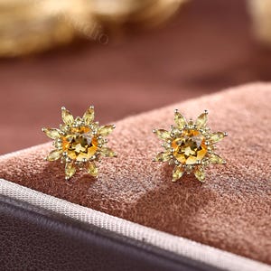 Può includere: Un paio di orecchini a lobo dorati. Ogni orecchino presenta una gemma gialla centrale, rotonda, circondata da gemme gialle più piccole a forma di petalo, creando un design a forma di raggio di sole. Gli orecchini sono esposti su una superficie marrone.