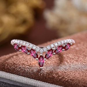 May include: A rose gold ring with a V-shaped design. The ring features a row of small, clear stones along the top and a series of marquise-cut, deep red gemstones in the center. The ring is set against a brown background.