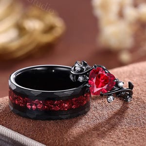 Può includere: Un anello nero con un intarsio di pietre preziose rosse e una gemma rossa a forma di cuore. L'anello presenta un motivo floreale con piccole gemme bianche. L'anello è esposto su una superficie marrone.