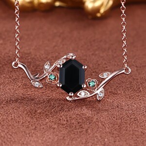 May include: Rose gold necklace featuring a black gemstone pendant. The pendant has an octagonal shape, accented with small green and clear gemstones, and a delicate leaf design. The necklace is displayed on a brown surface.