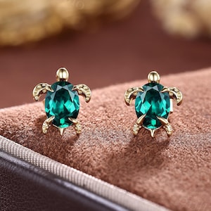 Könnte beinhalten: Ein Paar goldfarbene Schildkröten-Ohrstecker. Jeder Ohrring hat einen leuchtend grünen, oval geschliffenen Edelstein, der in einem schildkrötenförmigen Rahmen gefasst ist. Kopf und Flossen der Schildkröte sind mit goldenen Akzenten versehen.