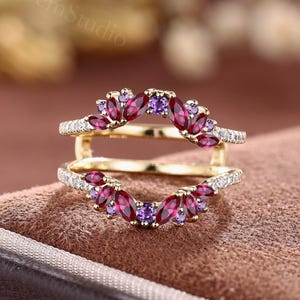 Könnte beinhalten: Ein goldener Ring-Guard bestehend aus zwei geschwungenen Bändern, die an den Seiten verbunden sind. Die Bänder sind mit tiefroten Steinen im Marquise-Schliff, kleinen runden violetten Steinen und winzigen klaren Pavé-Steinen am Rand besetzt.