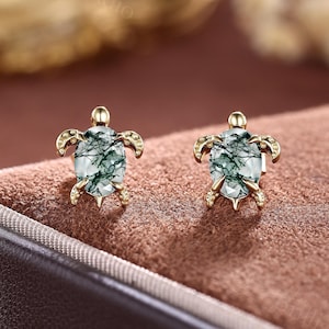 May include: A pair of gold-coloured turtle-shaped earrings. Each earring has a green and white gemstone body, with gold-coloured legs, head, and flipper details. The earrings are displayed on a brown surface.