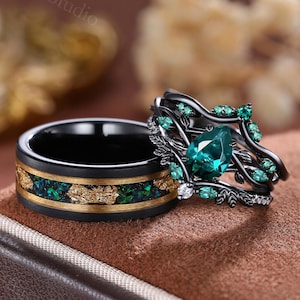 Puede incluir: Dos anillos: una banda negra con incrustaciones doradas y verdes, y un anillo negro con una piedra preciosa verde en forma de pera y pequeños acentos verdes. Los anillos se exhiben sobre una superficie marrón, mostrando su diseño.
